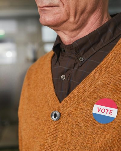 Gros plan d'un homme âgé en pull orange portant un badge "Vote" à l'intérieur.