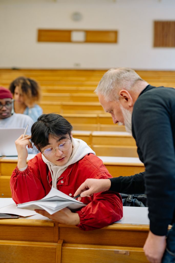 A teacher helps a student with coursework in a university lecture hall setting. Un enseignant aide un étudiant avec ses devoirs dans un cadre de salle de cours universitaire.