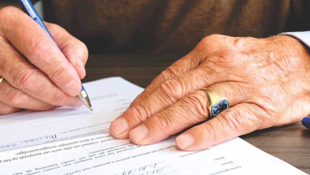 Close-up of a senior adult signing a legal document with a focus on hand and gold ring. Gros plan d'un adulte senior signant un document juridique avec un accent sur la main et la bague en or.