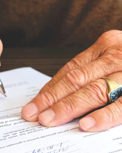 Close-up of a senior adult signing a legal document with a focus on hand and gold ring. Gros plan d'un adulte senior signant un document juridique avec un accent sur la main et la bague en or.