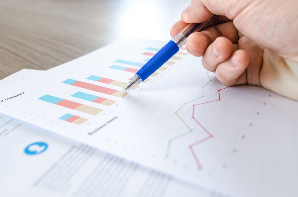 A close-up of a hand with a pen analyzing data on colorful bar and line charts on paper. Gros plan d'une main avec un stylo analysant des données sur des graphiques en barres et courbes colorés sur papier.