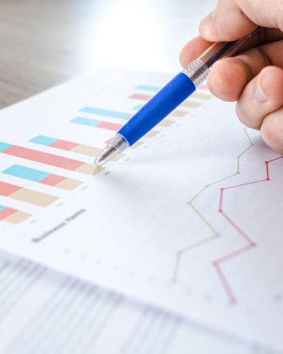 A close-up of a hand with a pen analyzing data on colorful bar and line charts on paper. Gros plan d'une main avec un stylo analysant des données sur des graphiques en barres et courbes colorés sur papier.